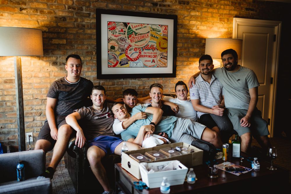 Group gathering on couch with pizza boxes and drinks in brick-walled room.