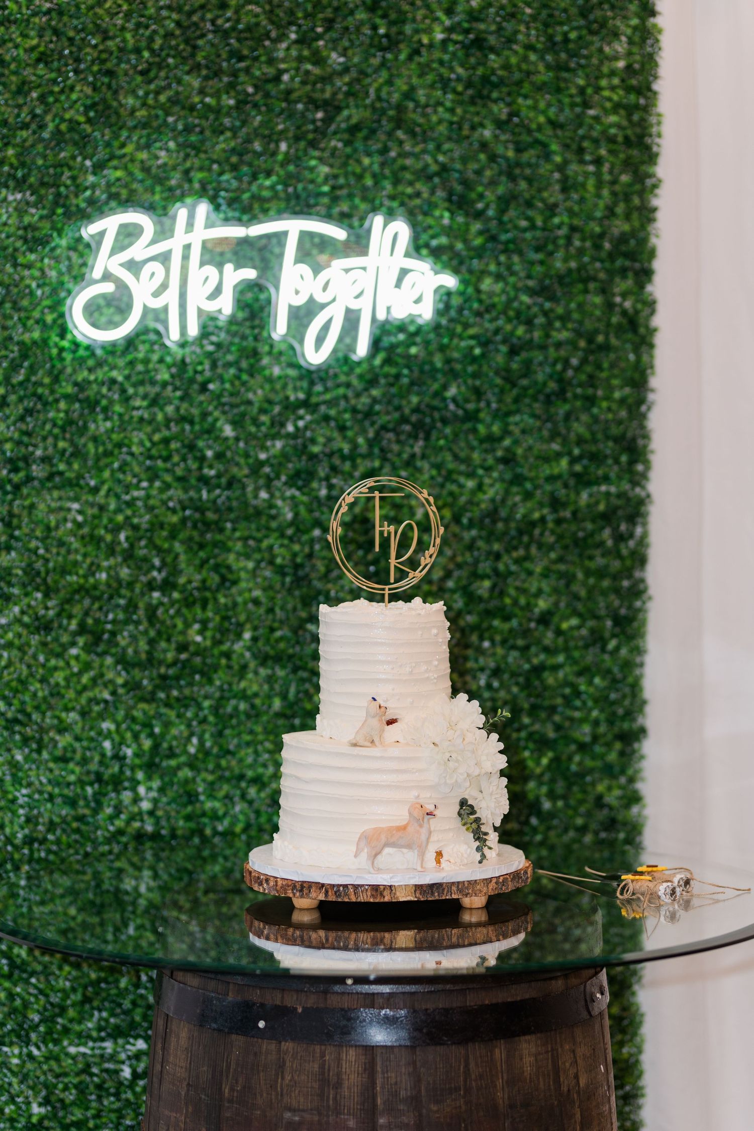 White wedding cake displayed on rustic wooden barrel stand against green hedge wall backdrop.