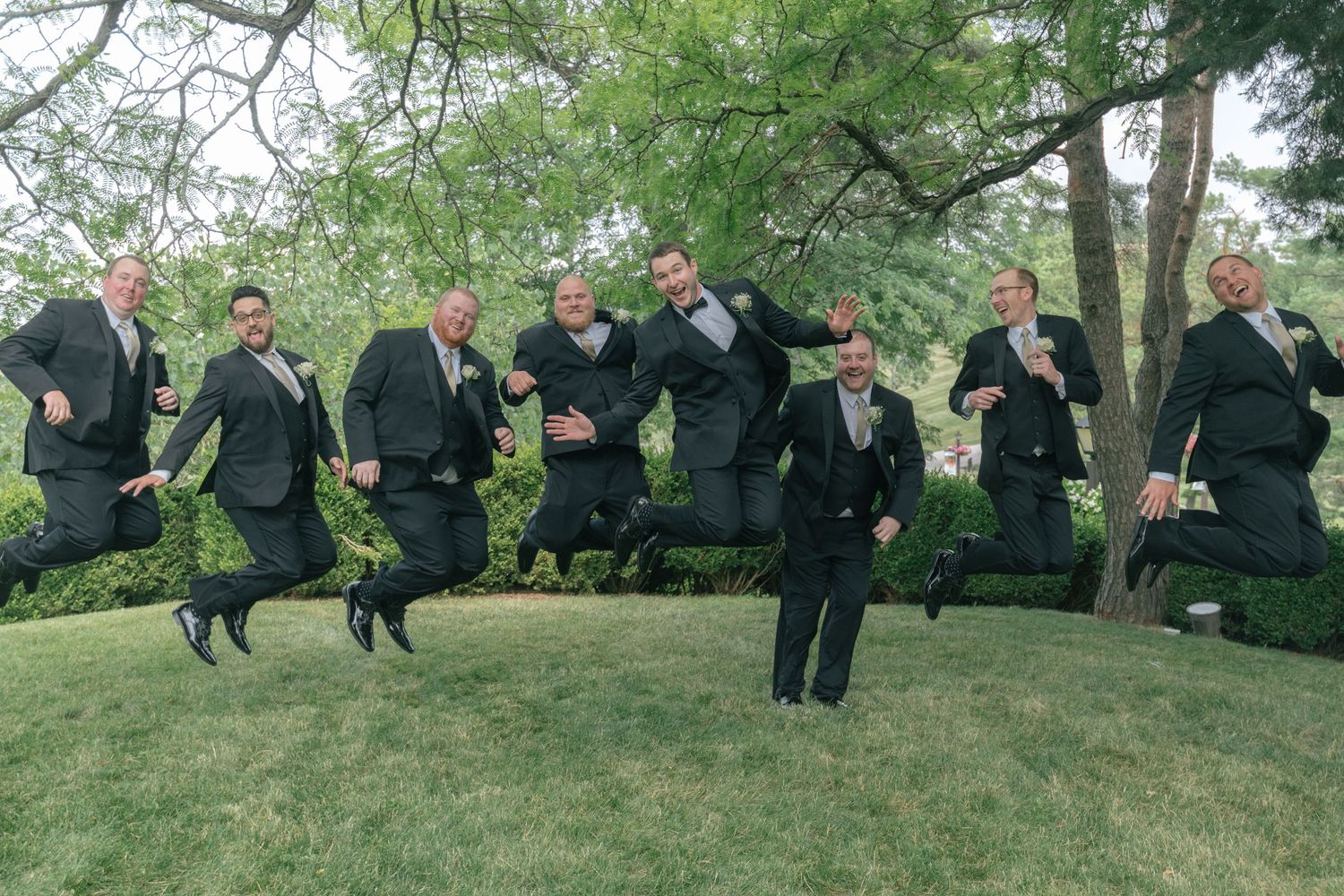 Group of men in dark suits jumping in unison on grass, one man stands still in center.