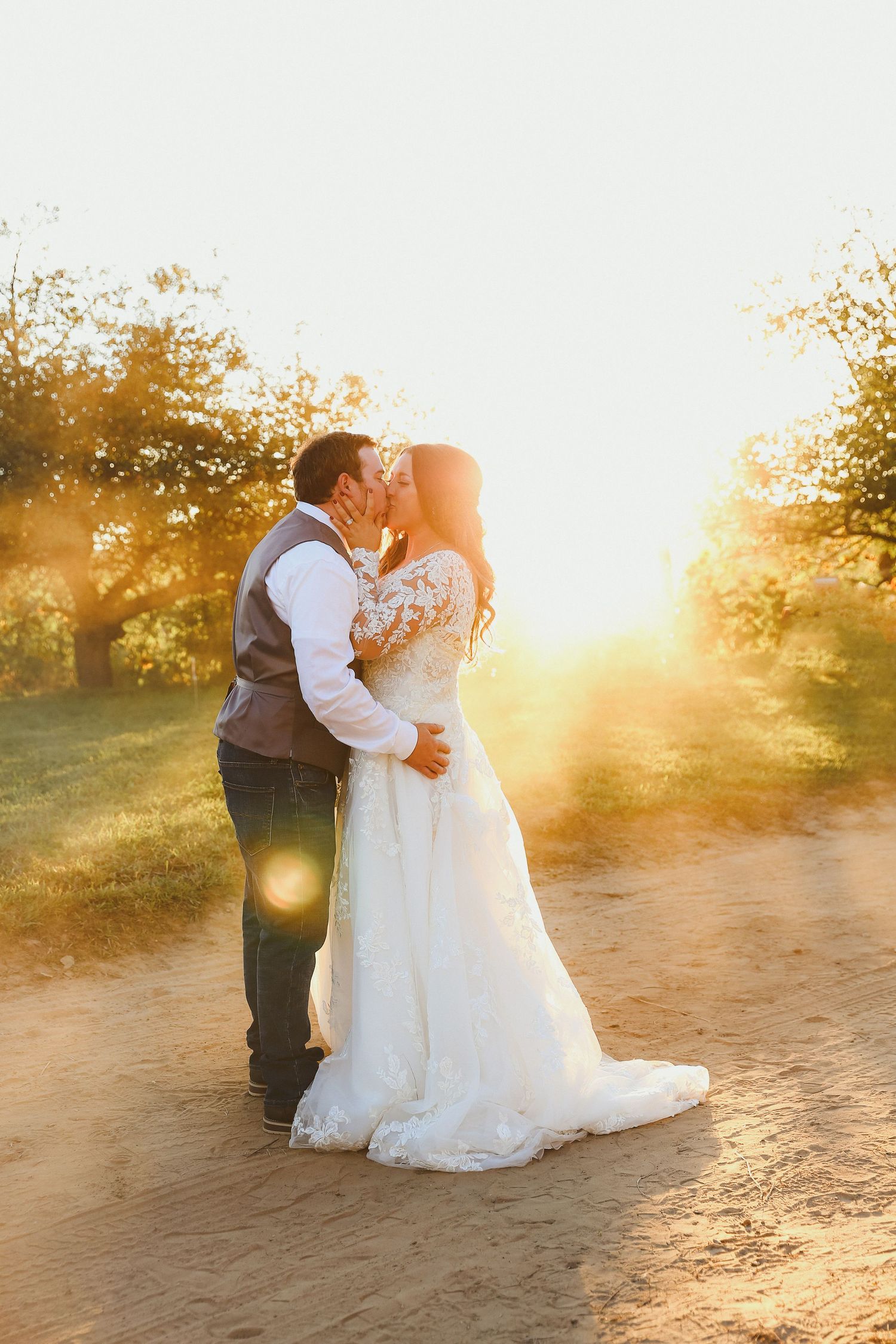 A couple in wedding attire share a romantic moment on a dirt path at sunset with golden sunlight streaming through trees.