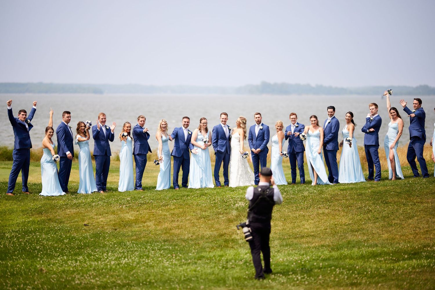 Large wedding party poses on waterfront lawn in blue suits and light blue bridesmaid dresses.