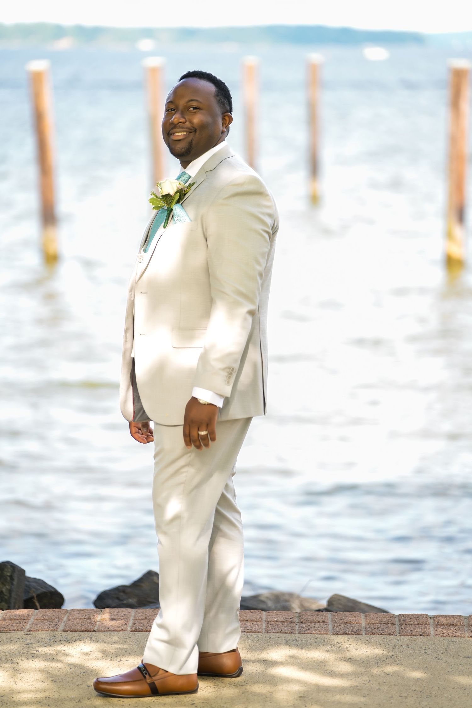 A person in a light beige suit poses by wooden dock posts near the water.
