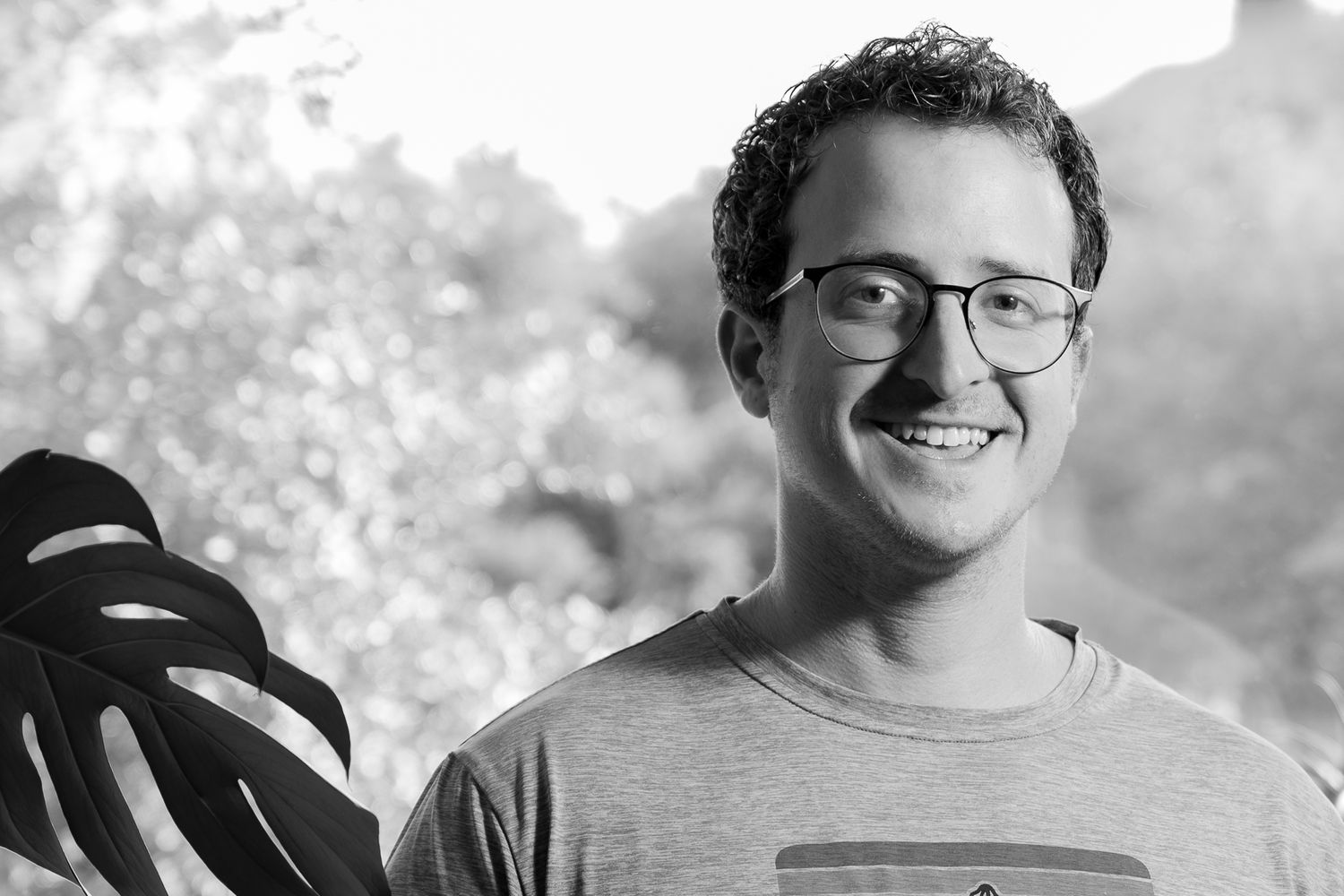 Portrait of someone in glasses and a gray t-shirt smiling against a bright outdoor background.