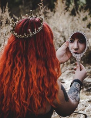 Woman with vibrant red wavy hair and gold crown holds ornate mirror reflecting her face outdoors.