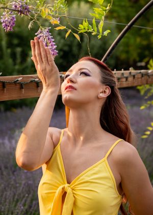 Beautiful redhead woman in yellow sundress reaching up to touch purple wisteria blooms outdoors.