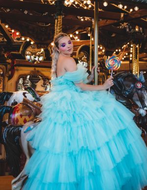 Woman in blue ruffled ball gown holding lollipop on ornate carousel with colorful horses.
