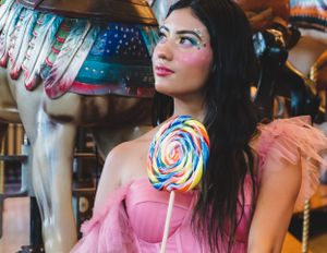 Woman in pink tulle dress holding rainbow lollipop beside carousel horse with artistic face makeup.