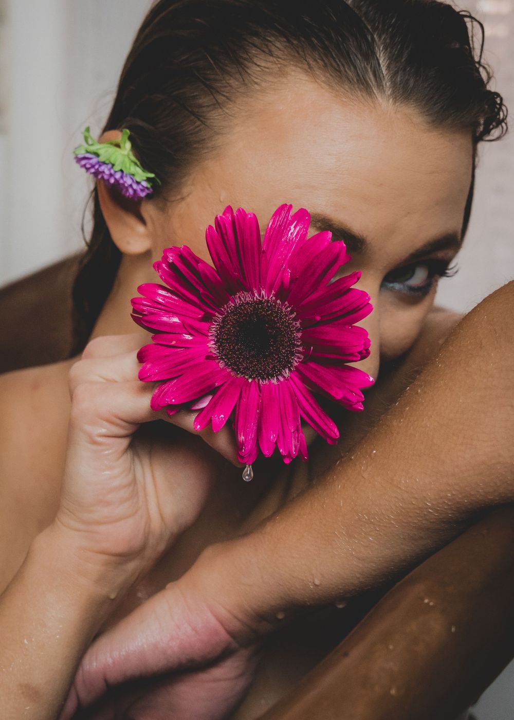 Close-up of woman holding pink gerbera daisy flower over her face with water droplets visible.