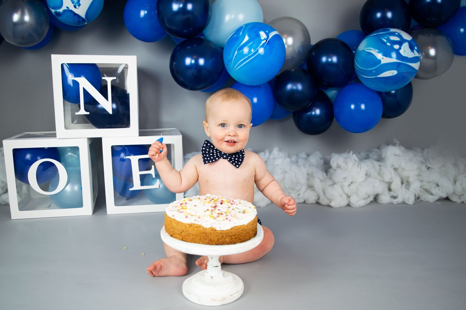 little boy eating cake for his first birthday on a grey backdrop, blue balloons by cake smash photographer in Doncaster