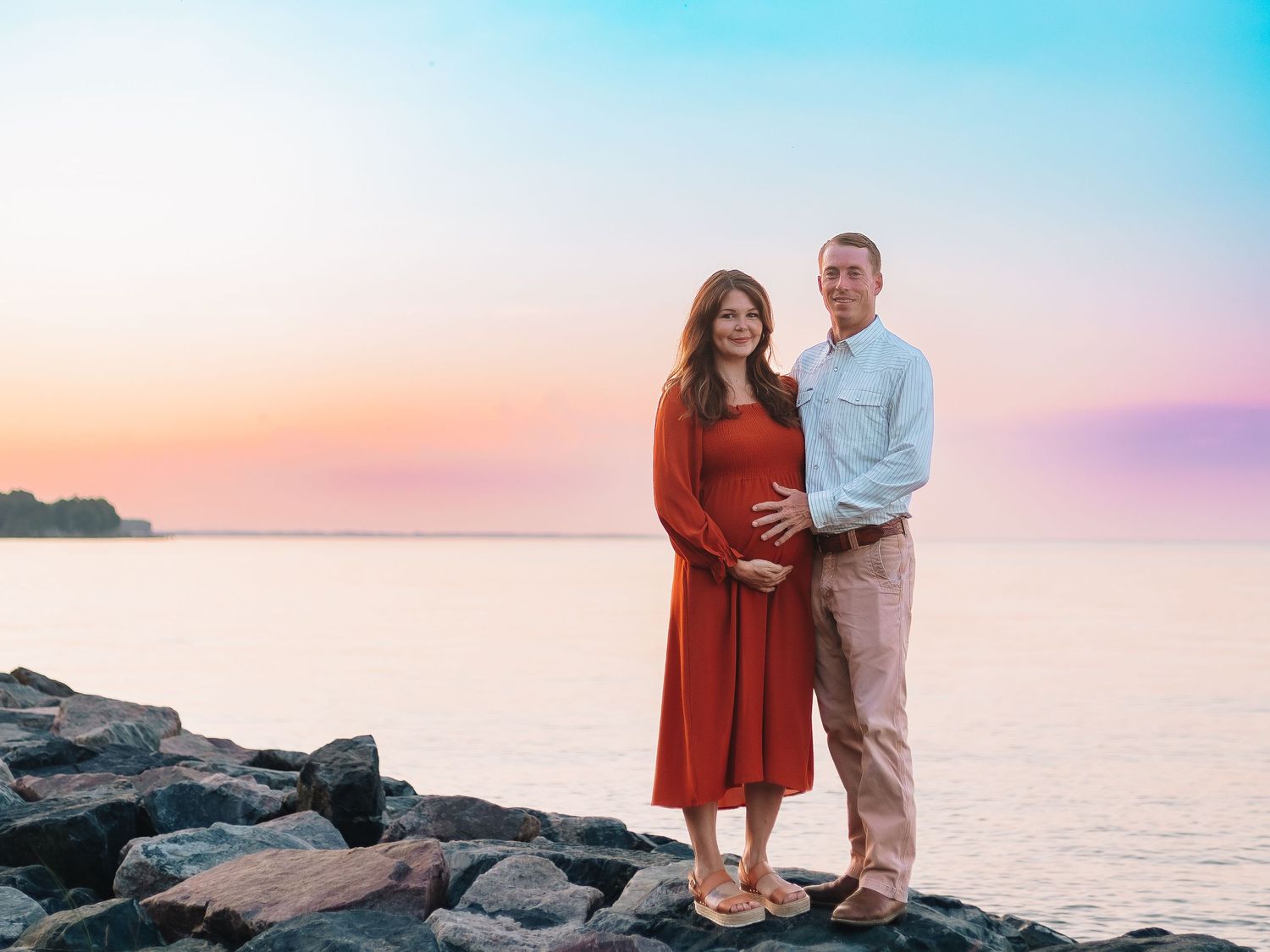 Romantic engagement photos on rocky coastline during colorful sunset.