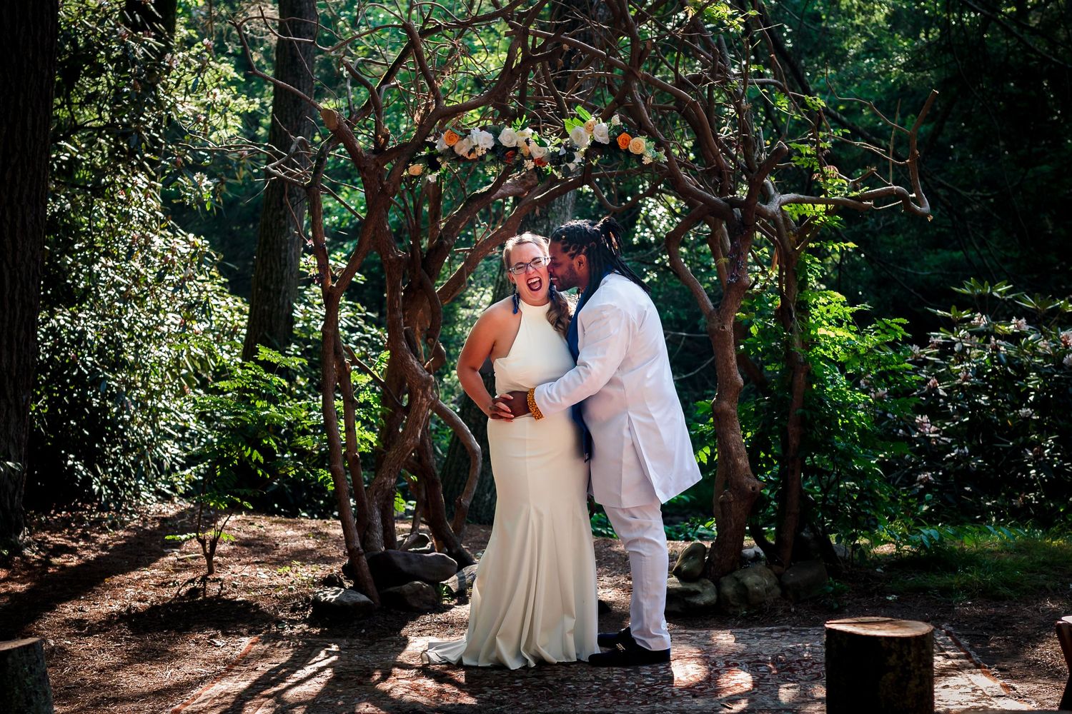 A couple shares an intimate moment under a rustic wooden arch adorned with flowers.