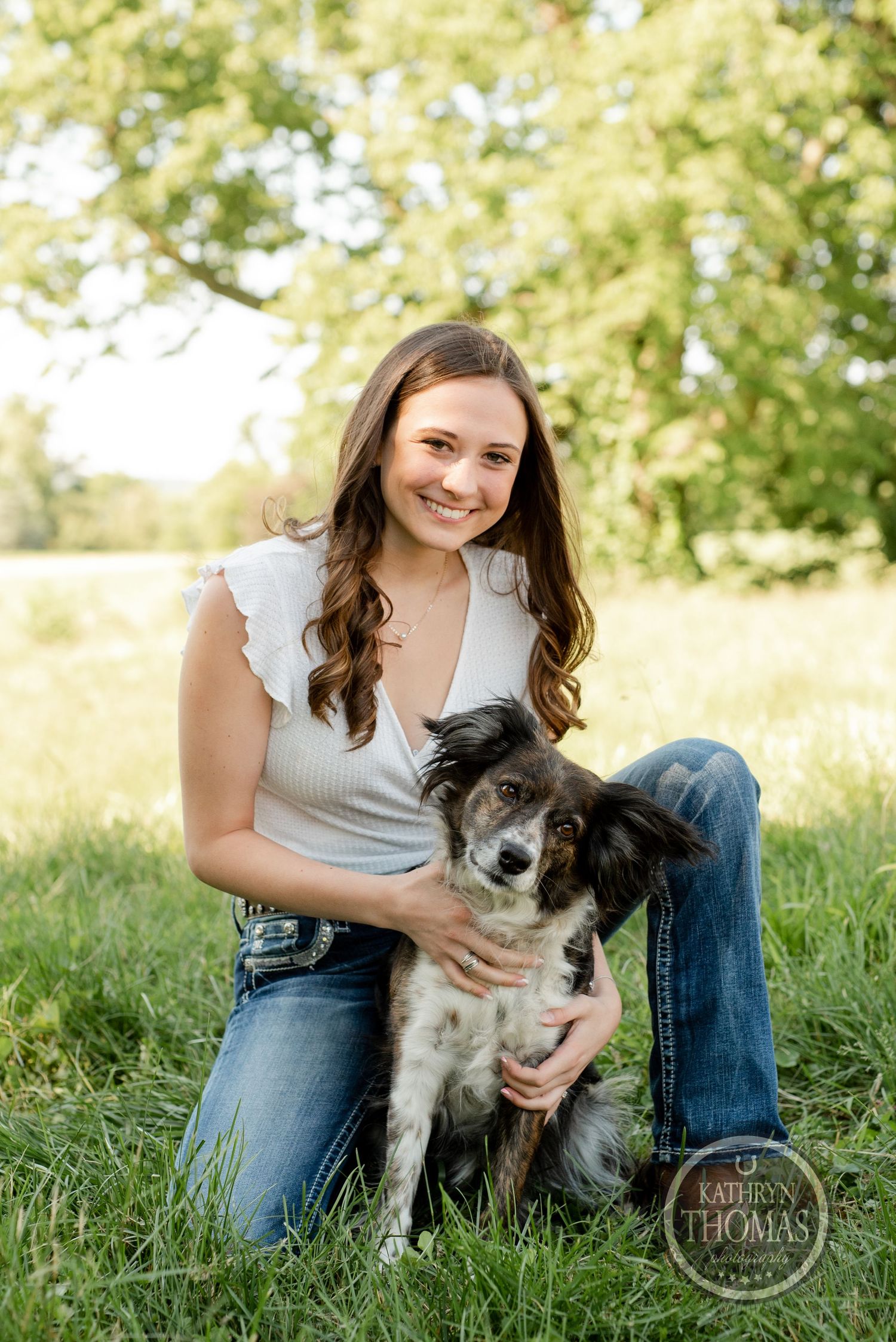 A woman in a white top sits with her black and white dog in a sunny meadow.