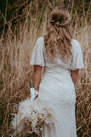 Bride in lace flutter-sleeve gown holding dried floral bouquet, standing in golden grass field with half-up floral hair.