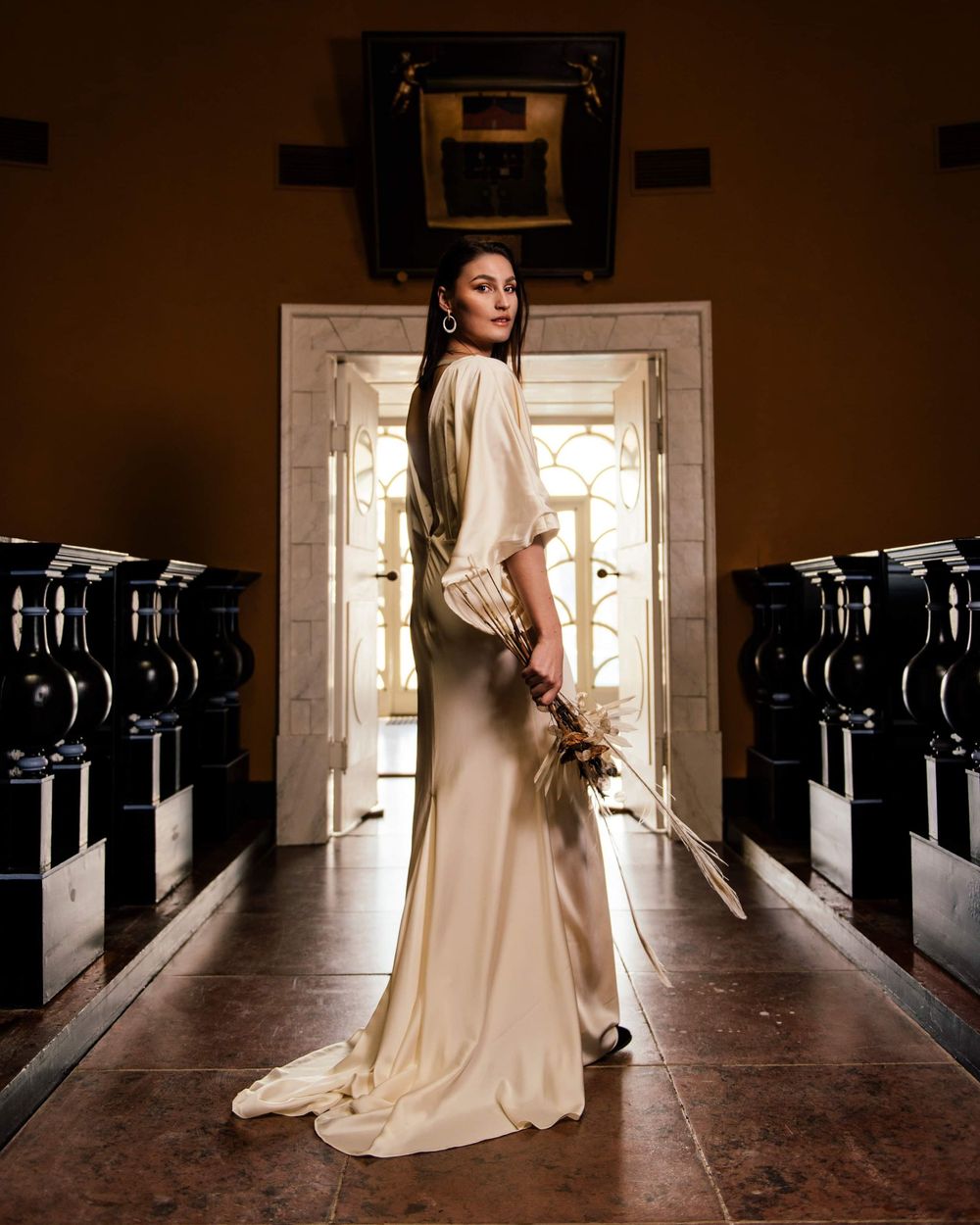 Elegant bride in a satin ivory gown holding a dried bouquet, posing in a grand hall with ornate doorways.