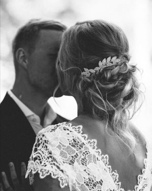 Black and white photo of a bride with lace dress and floral hair accessory, groom kissing her head at wedding.