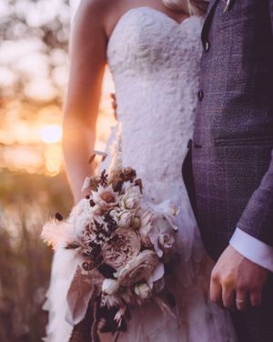 Bride in strapless lace gown holding blush floral bouquet beside groom in gray suit at golden sunset.