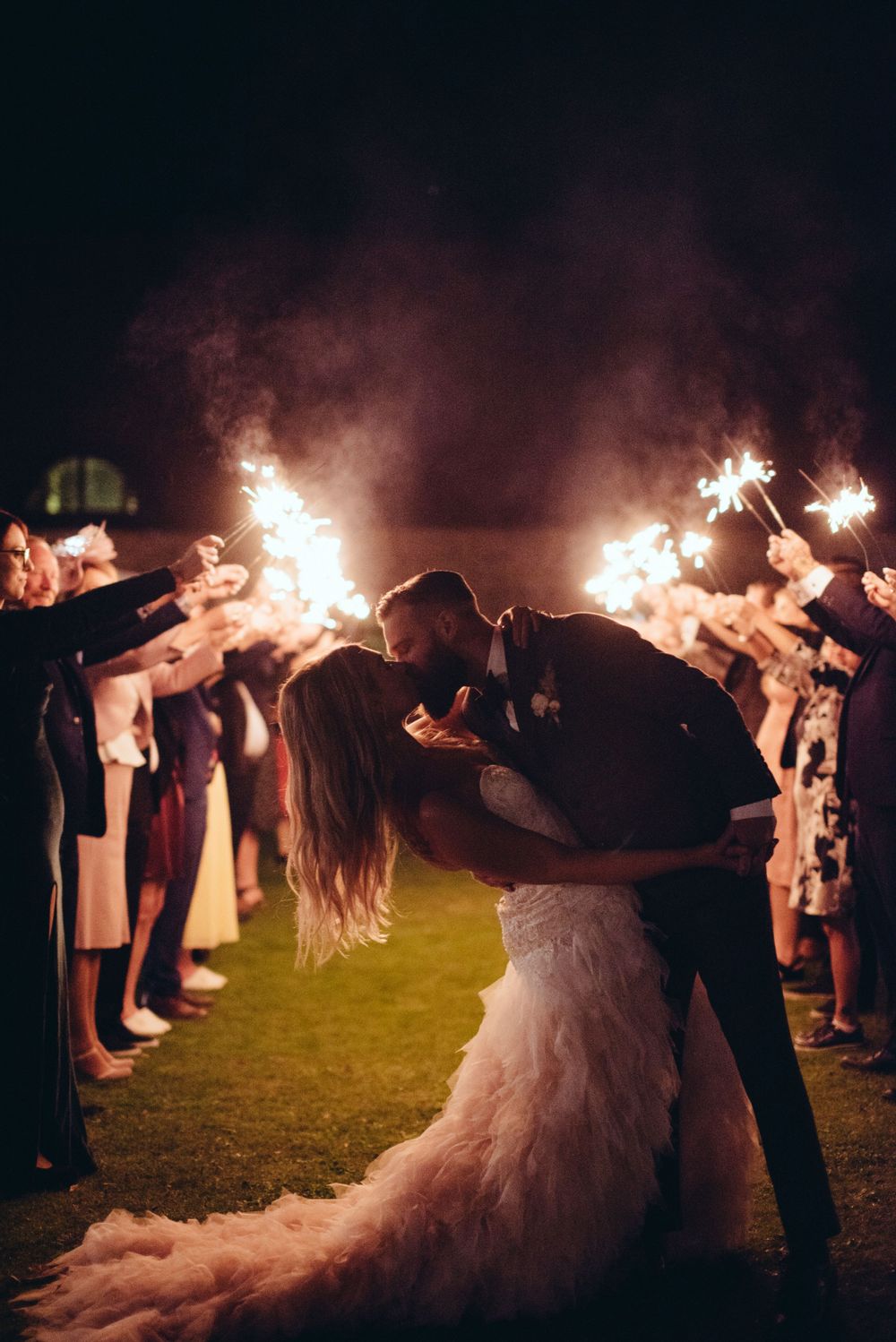 A romantic wedding couple shares a kiss under sparklers held by guests during their nighttime celebration on a lawn.