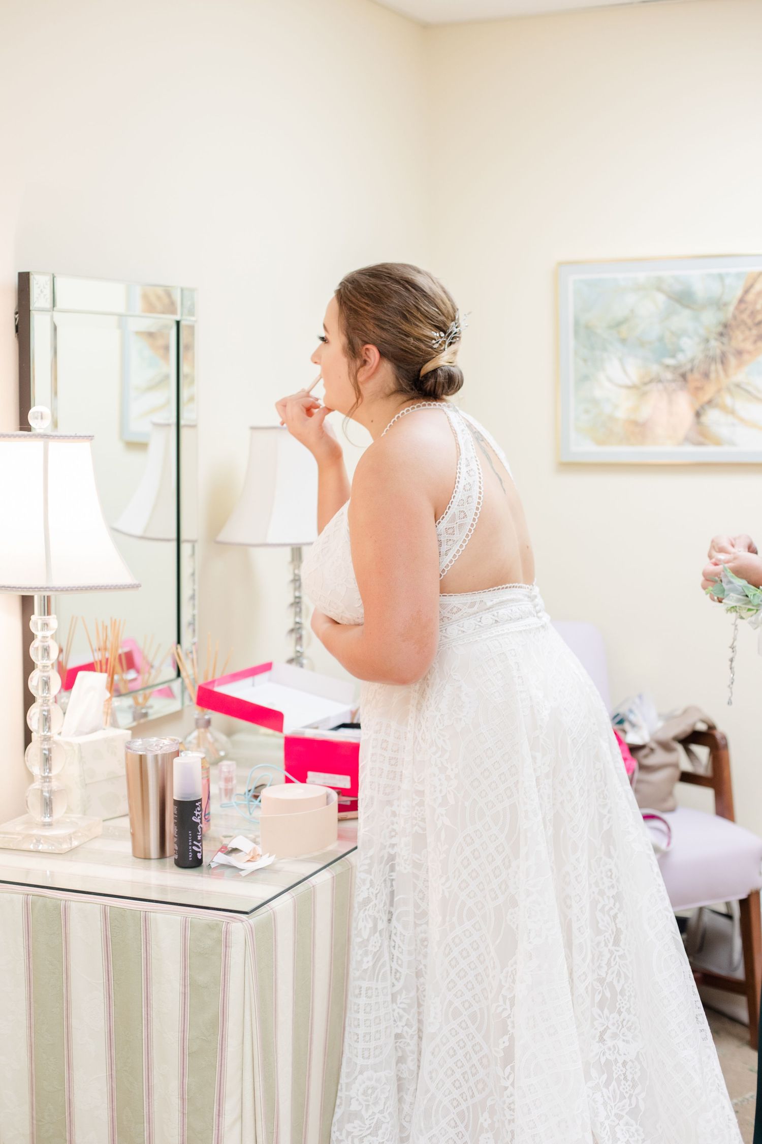 Bride in white lace dress getting ready in a bright room with vanity mirror.