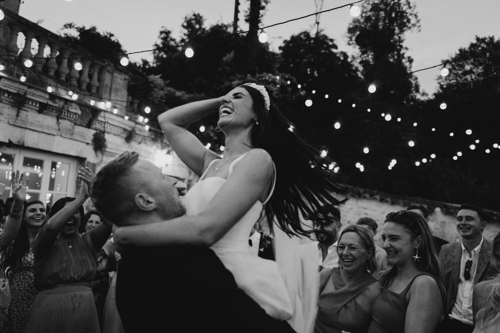 A couple shares a romantic moment under string lights at an evening wedding reception while guests look on in the background.