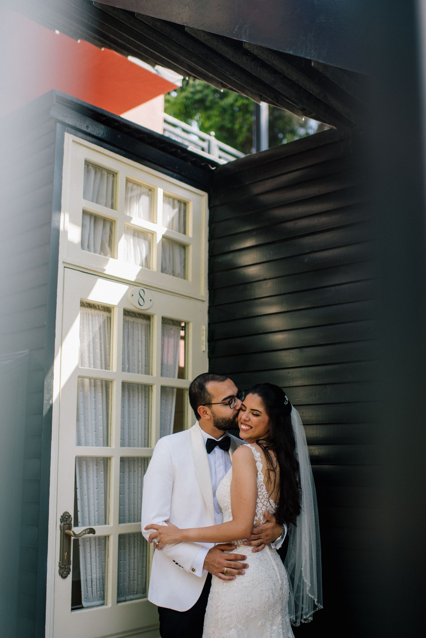 A couple shares an intimate moment against a white French door on their wedding day, with her in lace and him in white tuxedo.