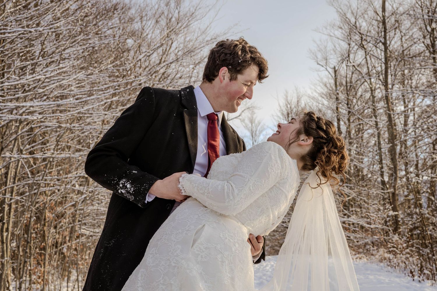 Wedding couple in winter scene shares romantic dip pose among bare trees.
