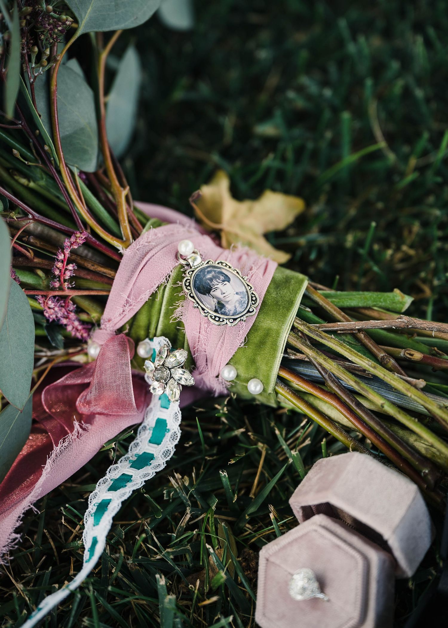 Wedding jewelry and rings arranged with pink ribbon and floral bouquet on grass.