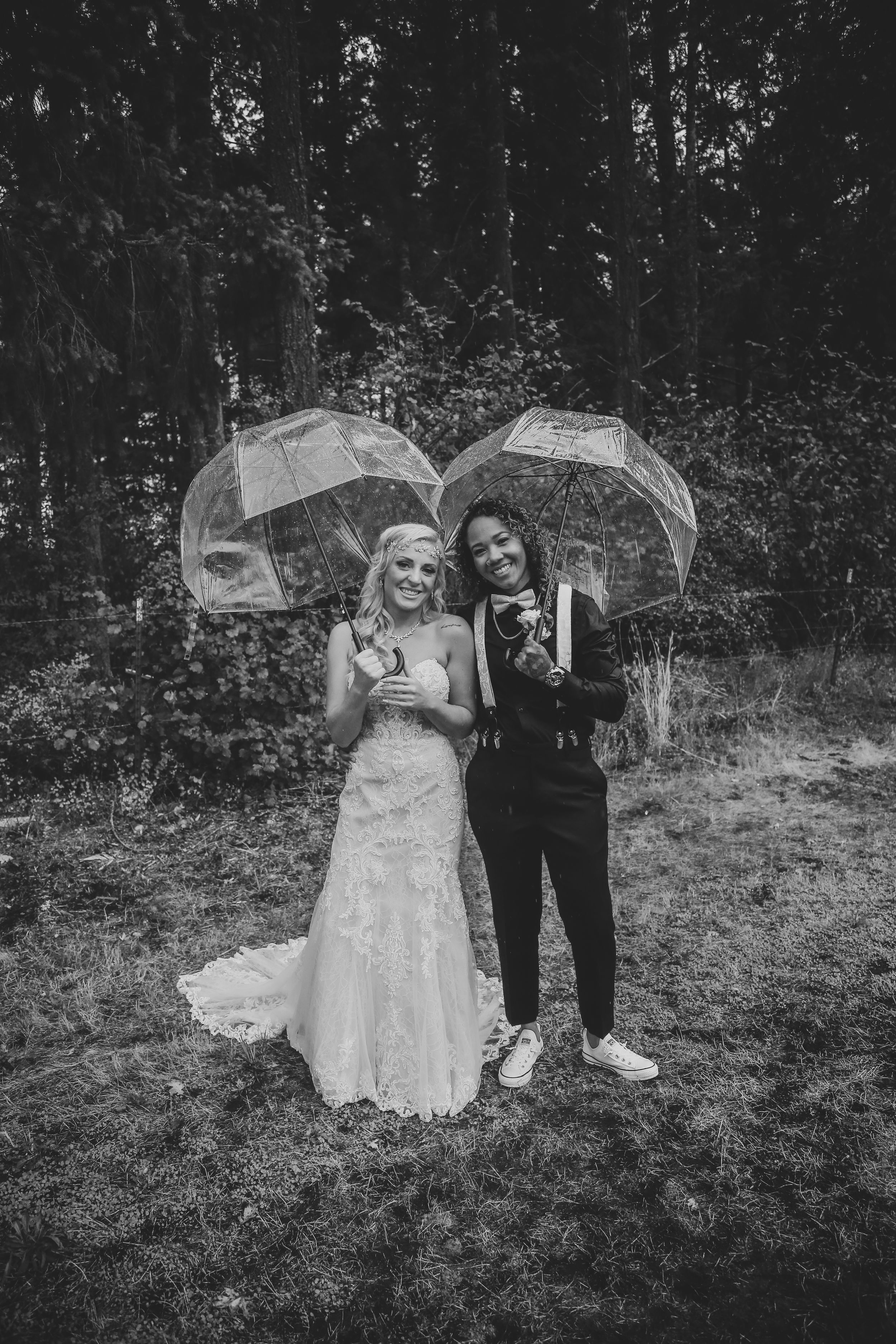 A black and white wedding photo of a couple with clear umbrellas in a forest setting.
