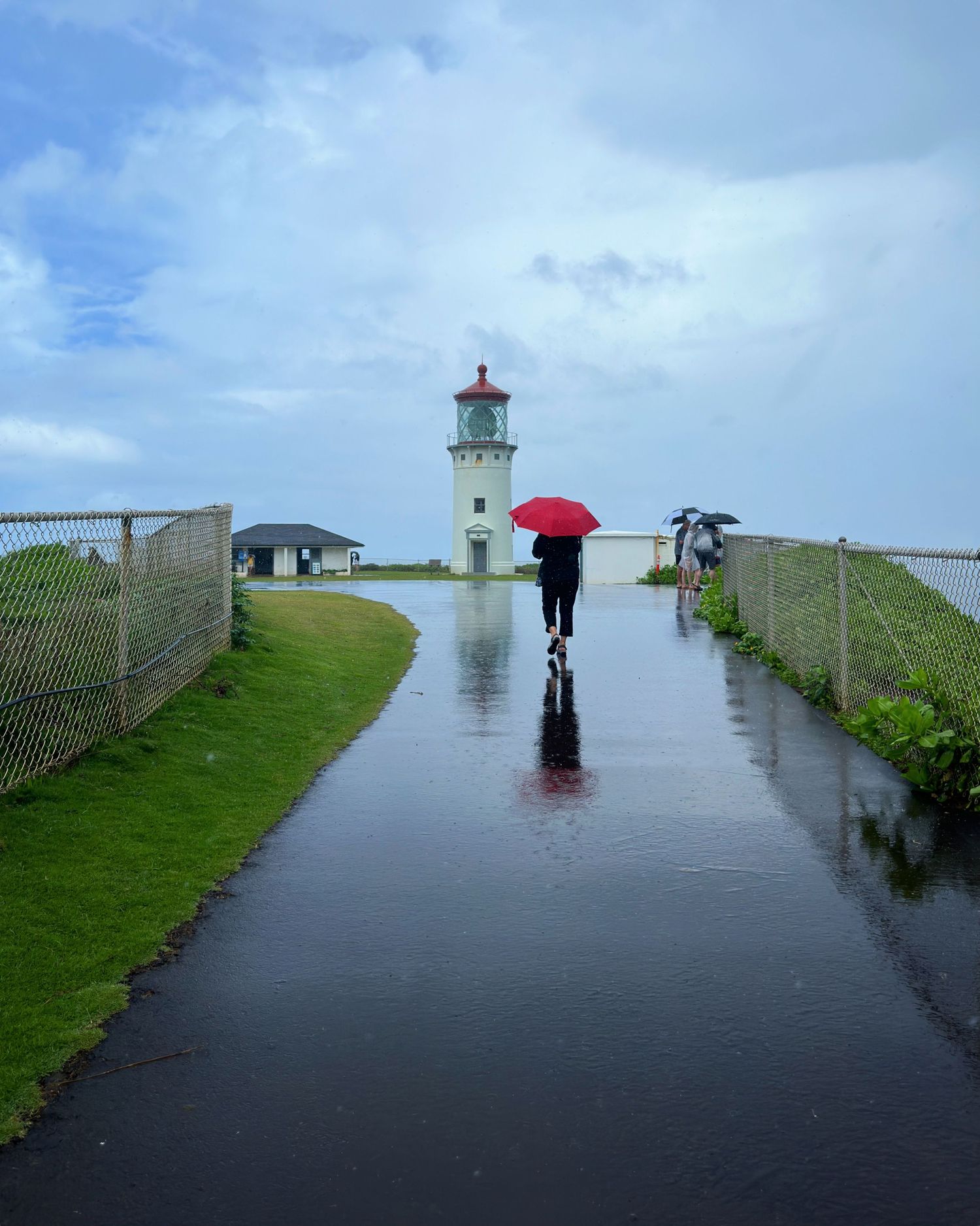 Person walking toward a lighthouse in the rain carrying a red umbrella on a wet path.