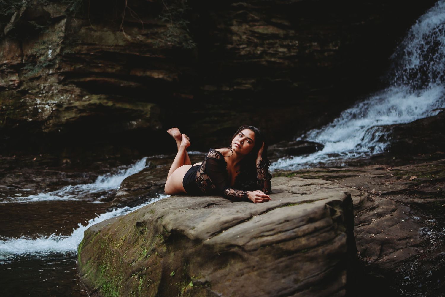 Figure reclining on rocks near cascading waterfall in natural setting.