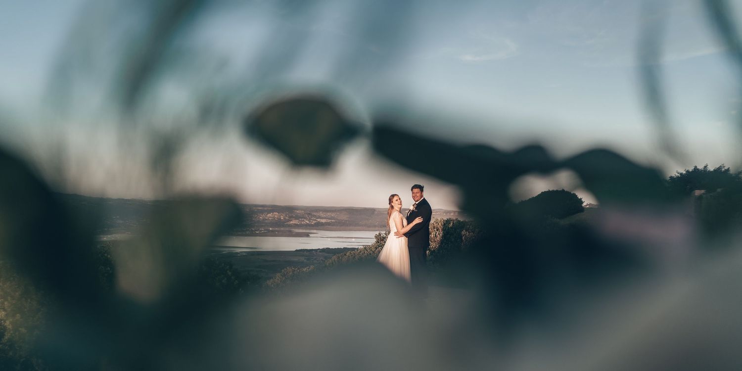 Wedding couple standing close together framed by blurred leaves with ocean backdrop at dusk.
