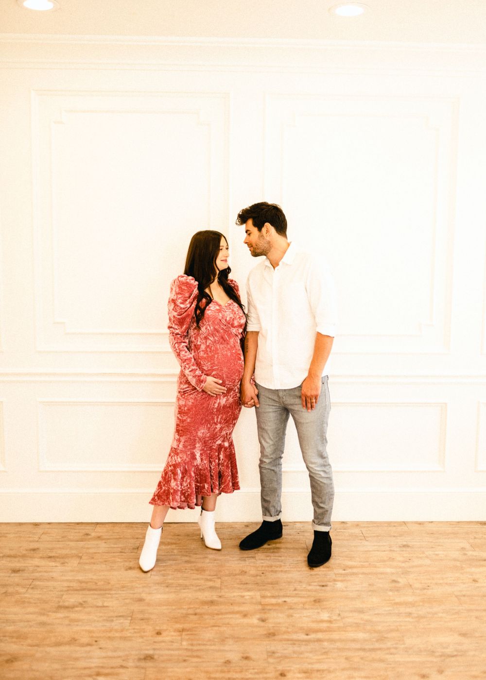 Maternity photography session with couple in red dress and casual wear on wooden floor.