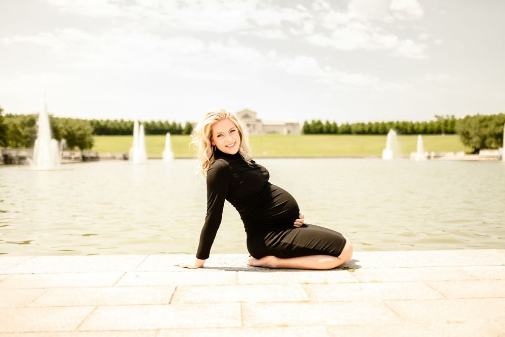 A maternity photo shoot by a lake with fountains in the background on a sunny day.