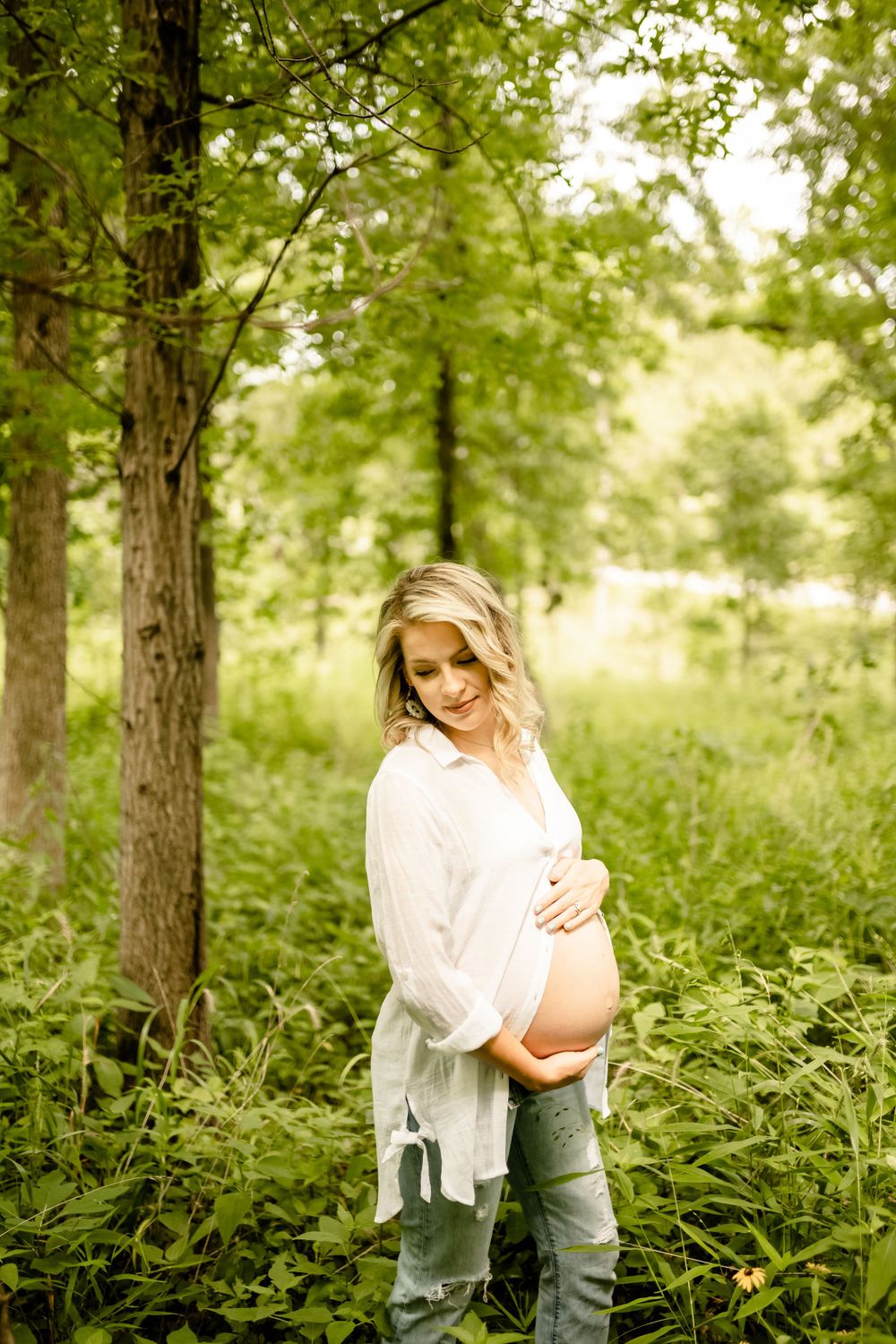 Maternity photo of woman in white shirt among lush green forest setting.