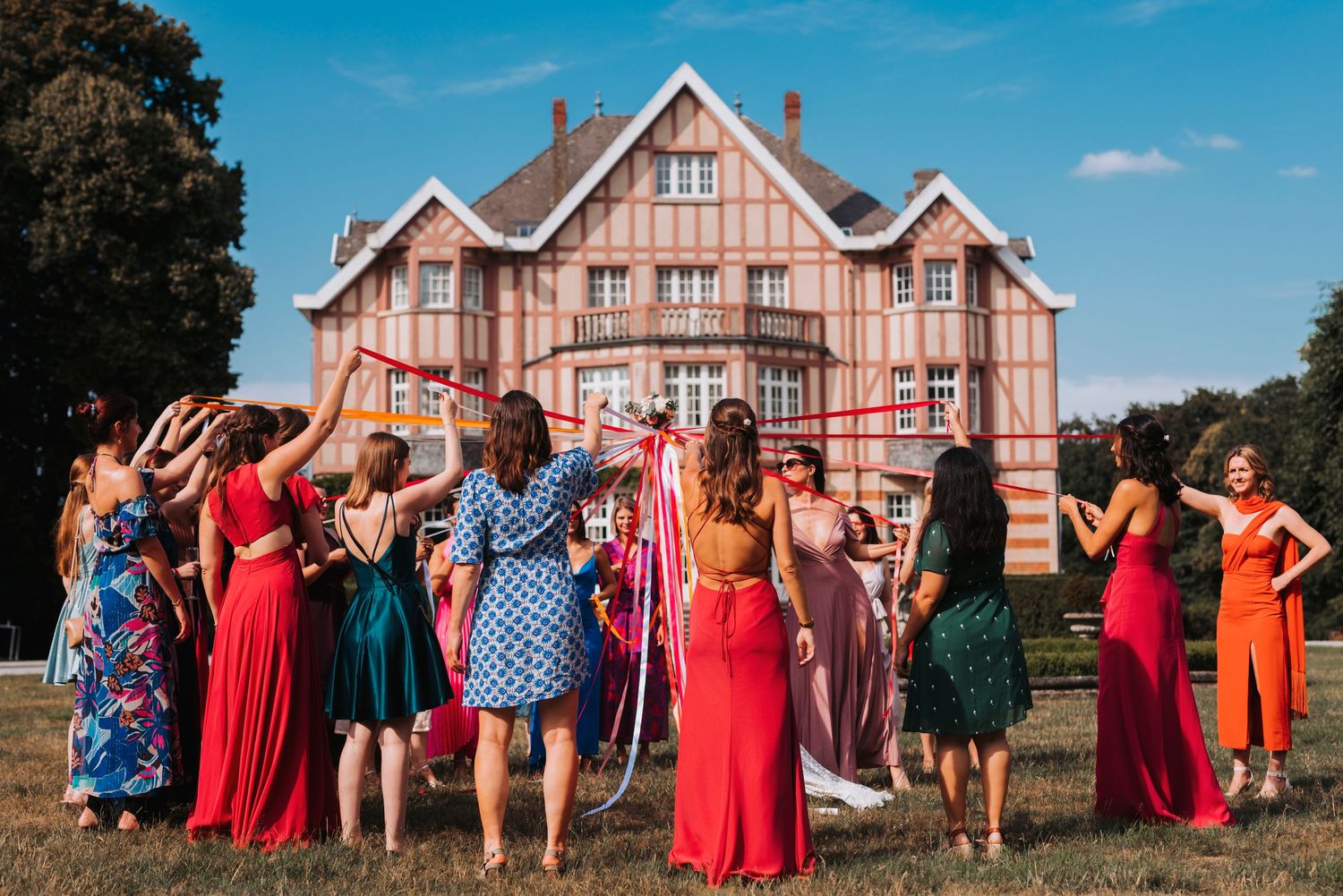 Celebratory group in colorful dresses gather outside a grand wooden mansion on a sunny day.