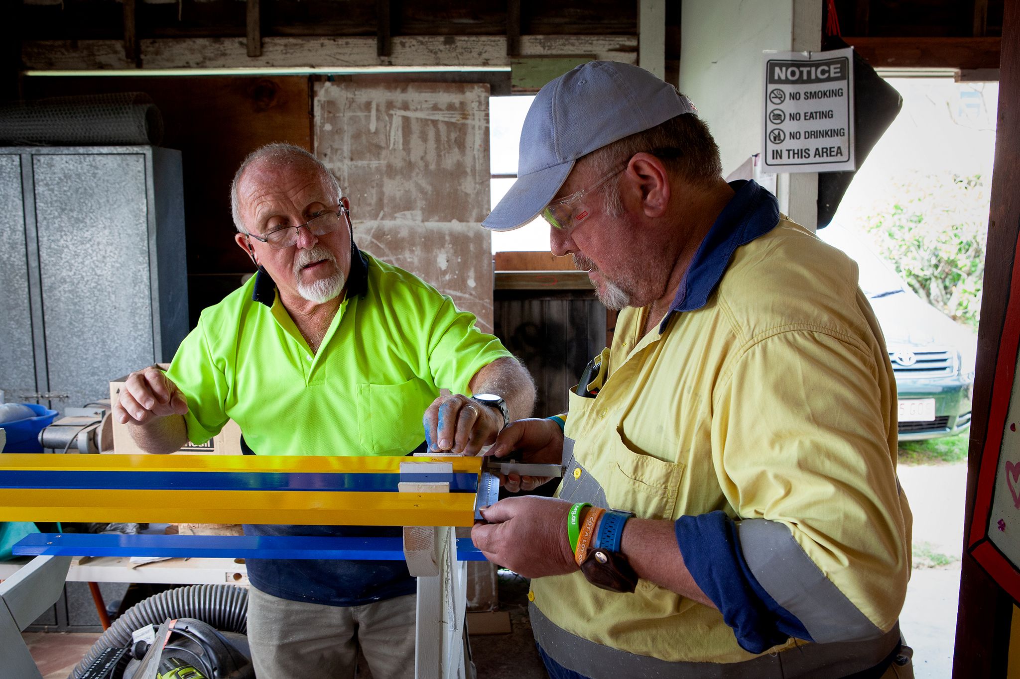 Two middle-aged men work on a wooden table in a workshop.