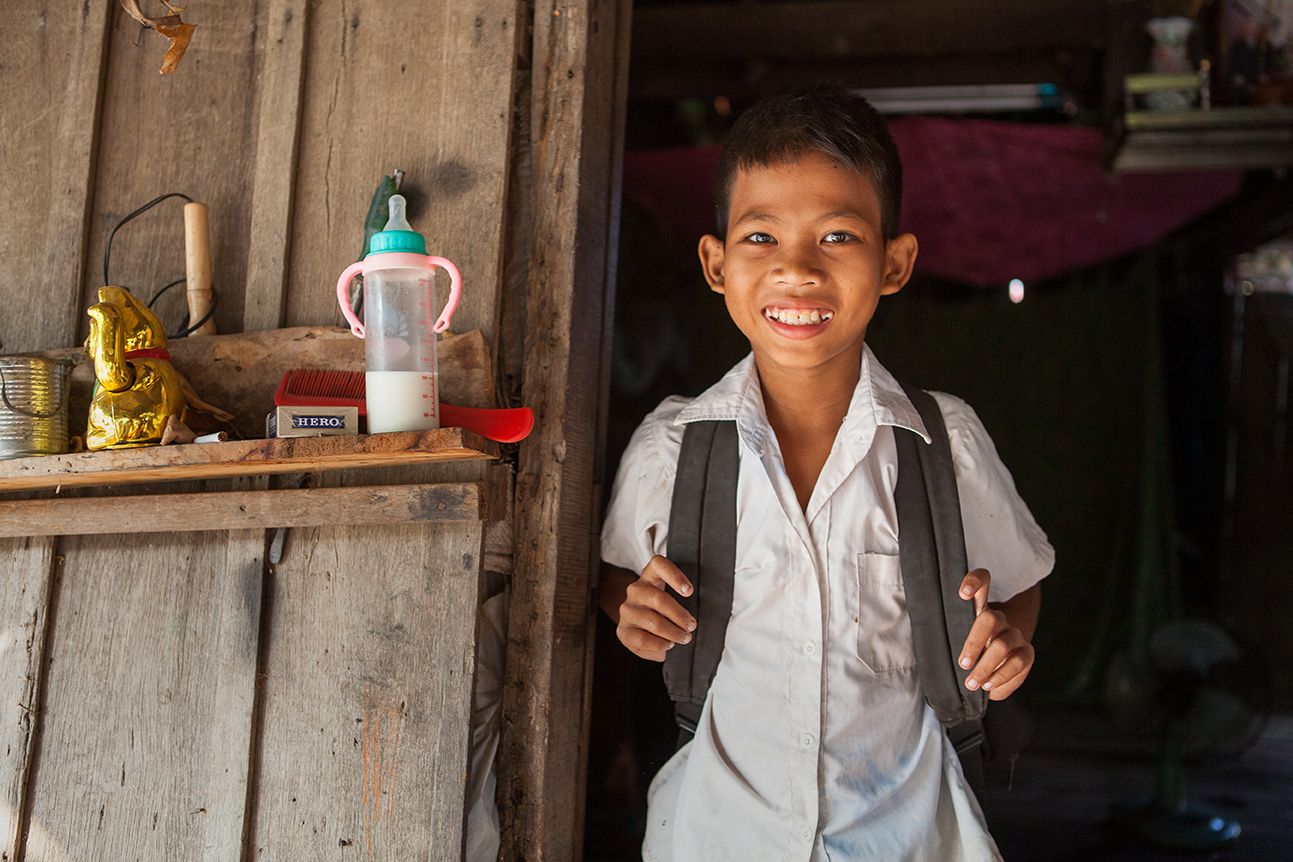 A young Cambodian boy wearing a school uniform stands in a doorway smiling to camera.