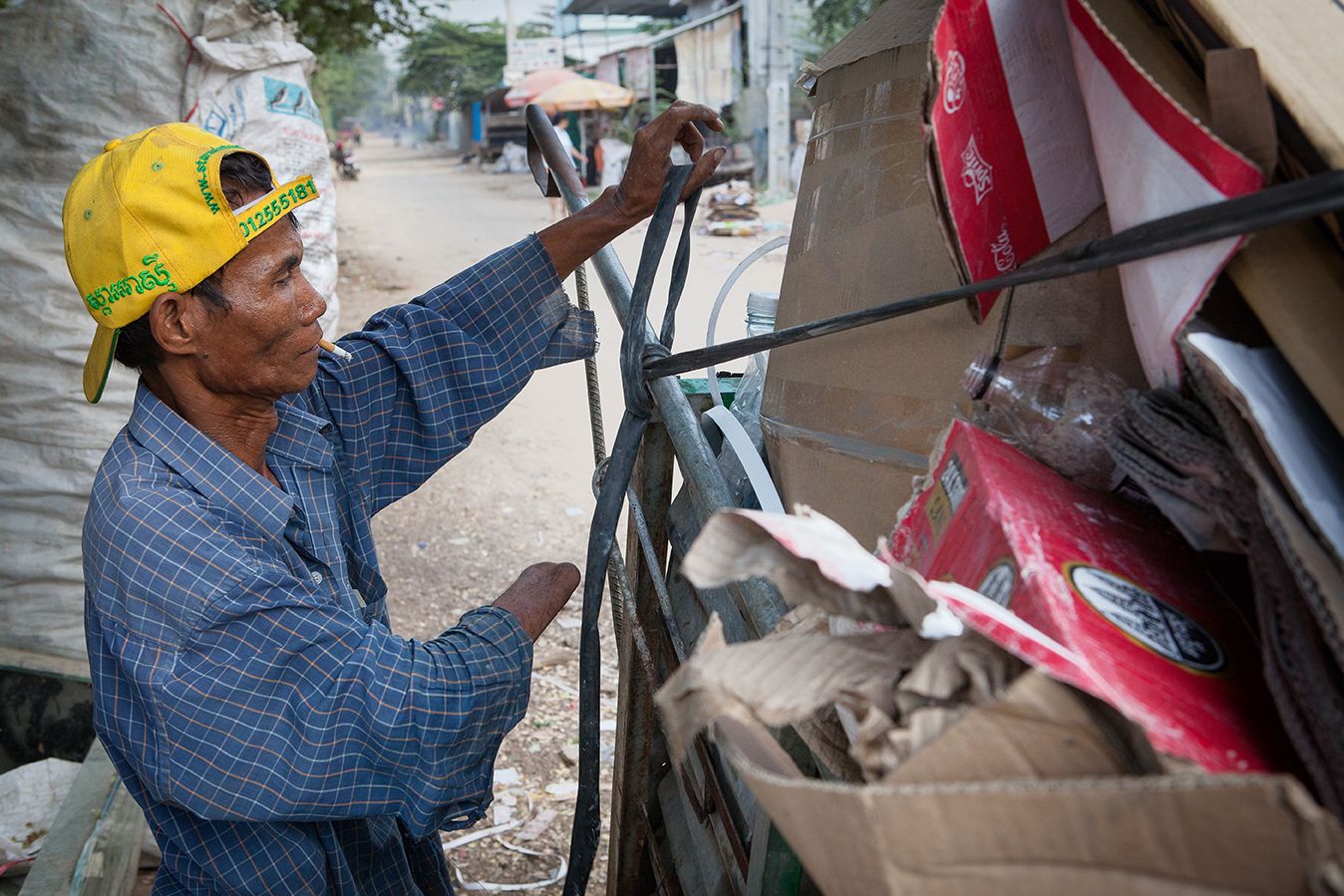 A Cambodian man with only one hand ties up a trailer of recycling as he smokes a cigarette.
