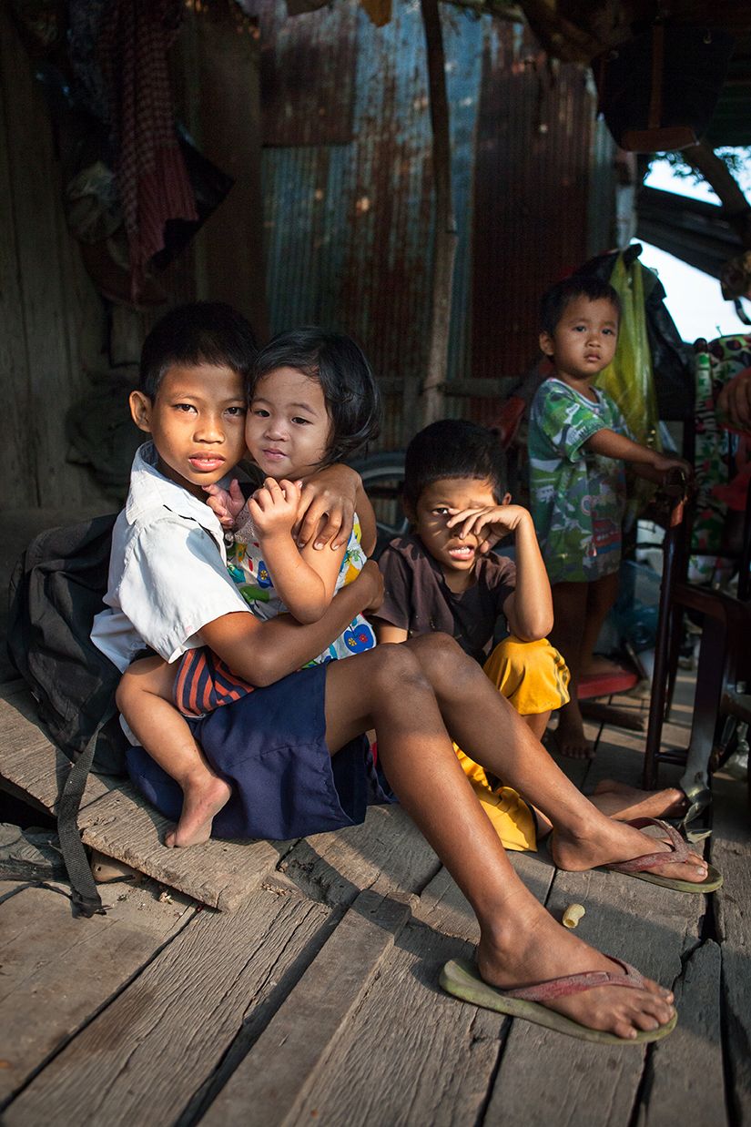 A group of young children sit on the balcony of a shack in a low socioeconomic area, all looking to camera.
