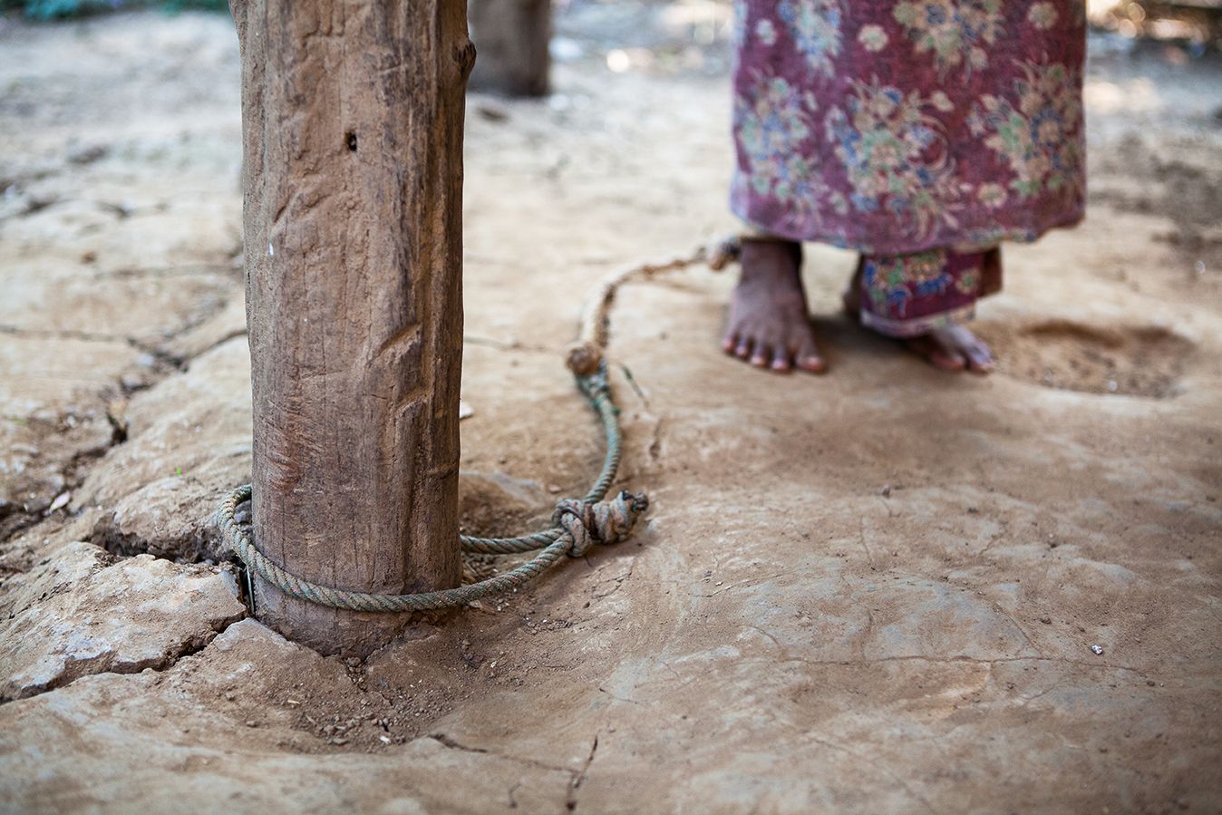A rope tied around a wooden pole leads the the foot of a female figure in the background who is tied to it.