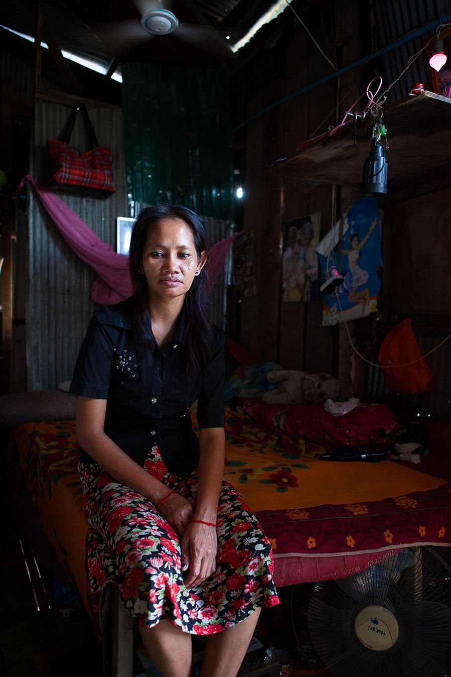 A middle-aged Cambodian woman sits on the edge of a bed in a dark shack.