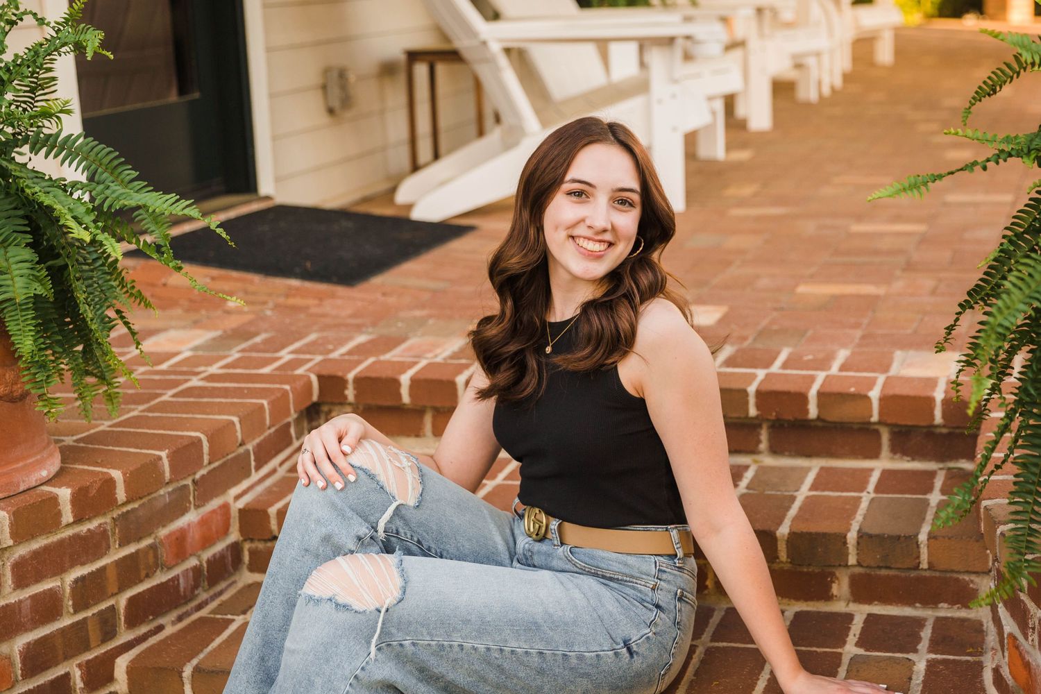 Senior girl in black top and jeans sitting on brick steps with hanging plants.