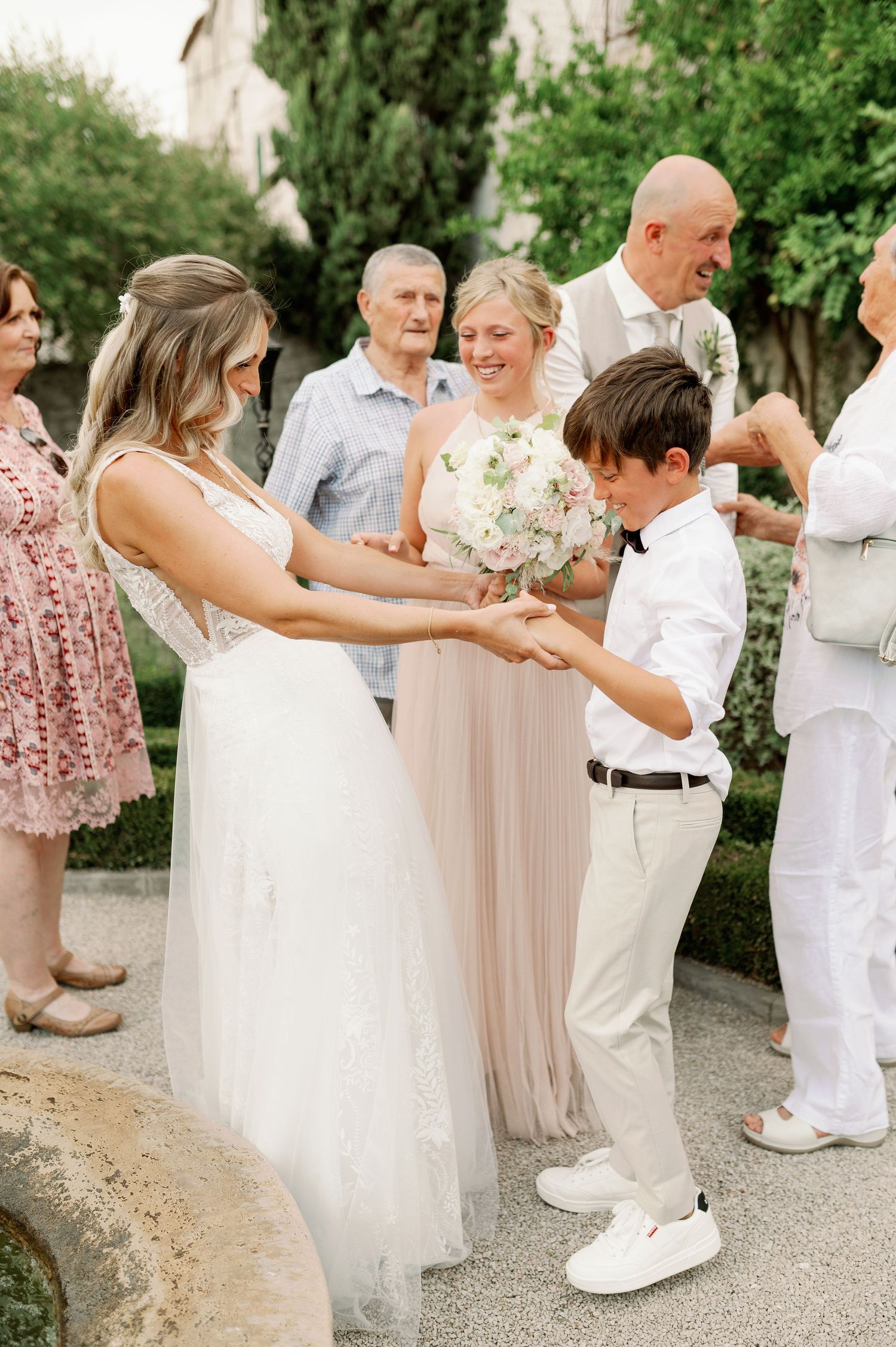 Wedding celebration in garden setting with guests gathering around bride in white gown.