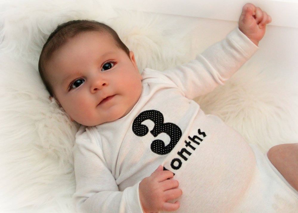 Baby in white '3 month' onesie lying on soft white blanket with arm raised.