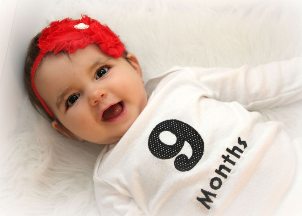 Baby wearing 9 month onesie and red headband smiling against white background.