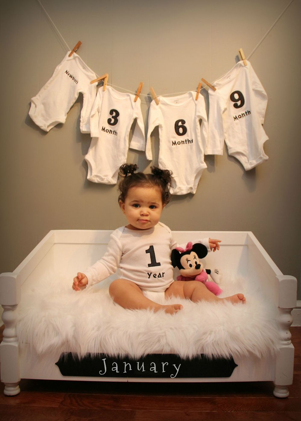 Monthly milestone photo with white onesies hanging above and soft white bench with Mickey Mouse plush toy.
