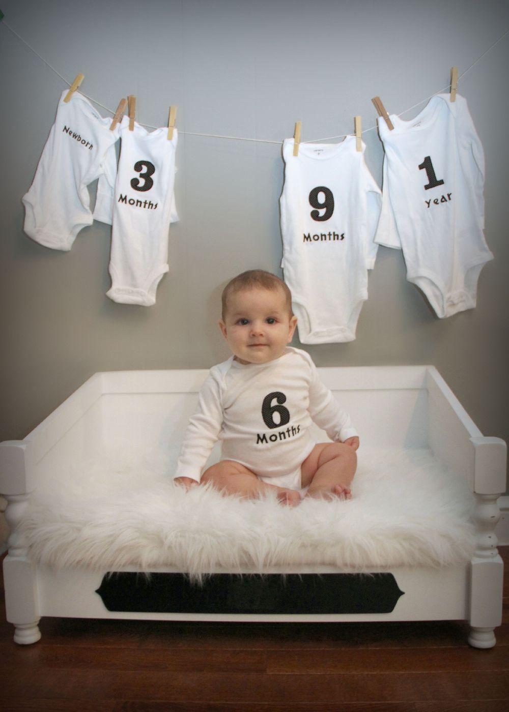 Monthly milestone photo with numbered onesies displayed on clothesline above white bench.