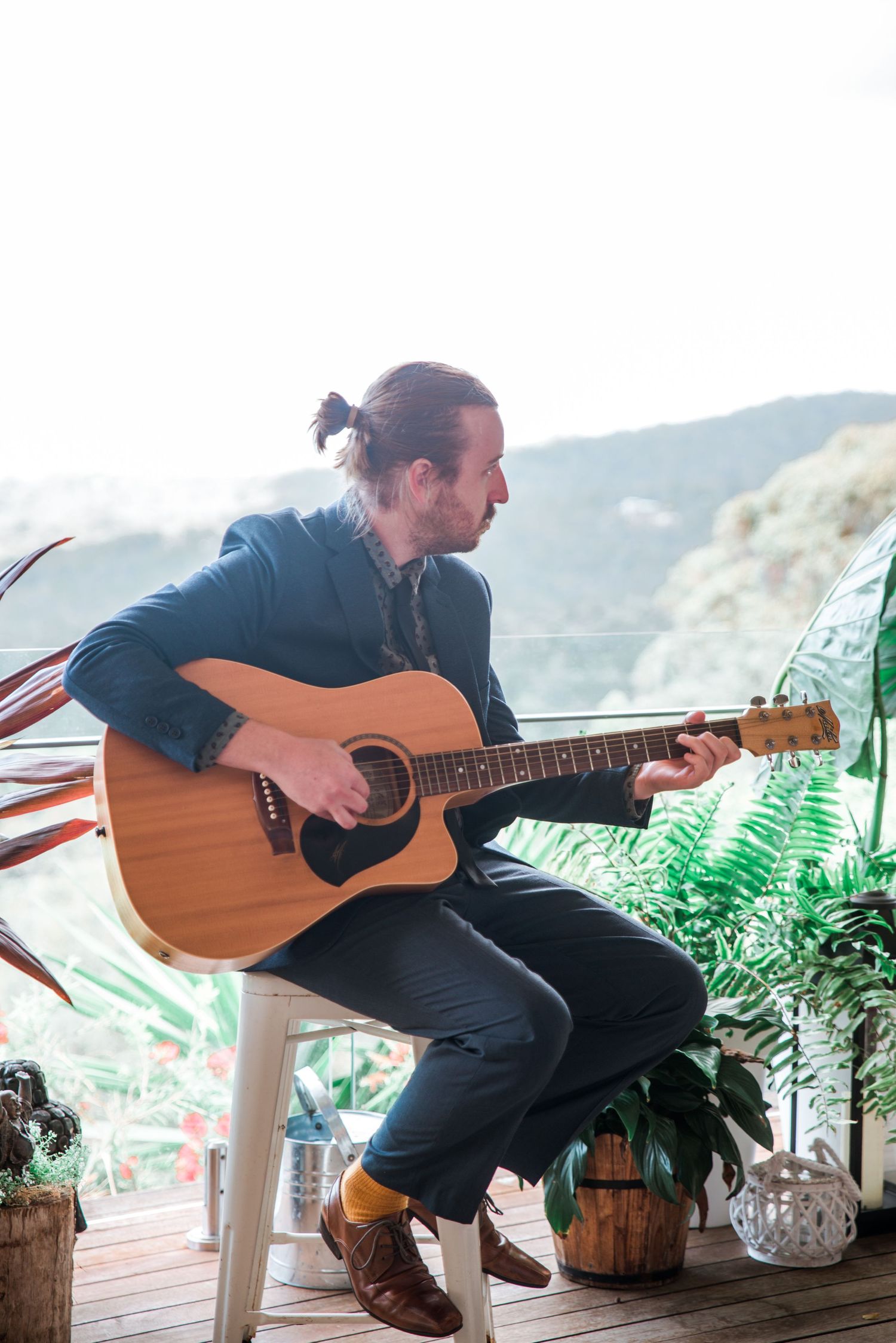 Musician playing acoustic guitar on covered porch with mountain views and potted plants.