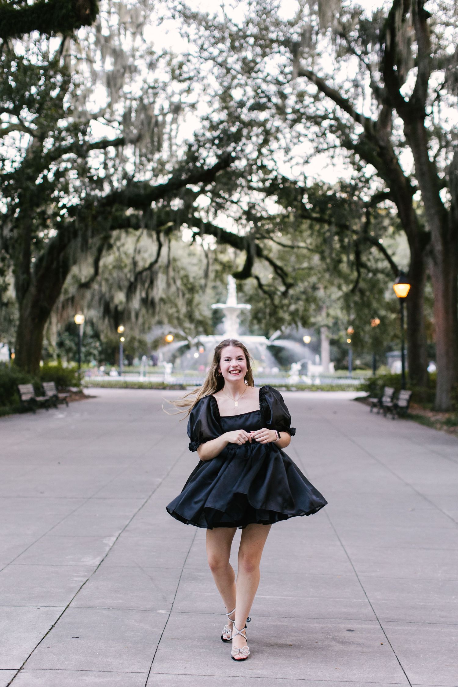 A person twirling in a black dress on a tree-lined path with a fountain in the background.