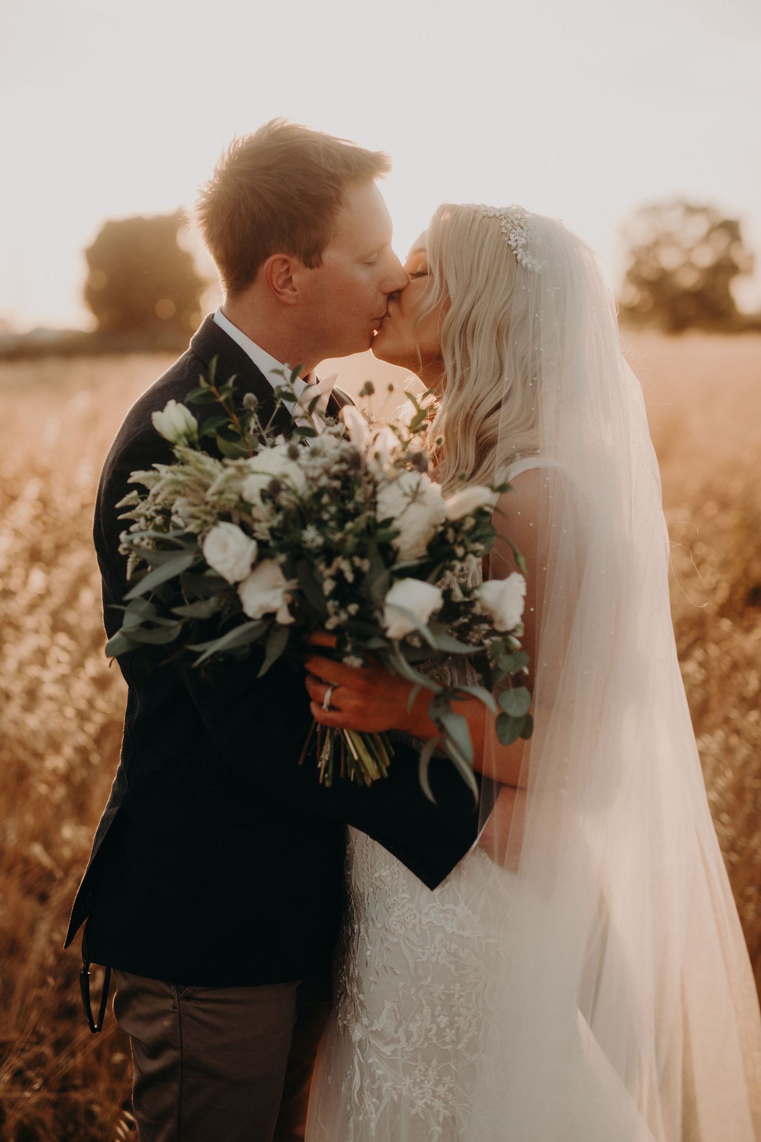 Romantic wedding couple sharing a kiss at sunset in a golden field with floral bouquet.