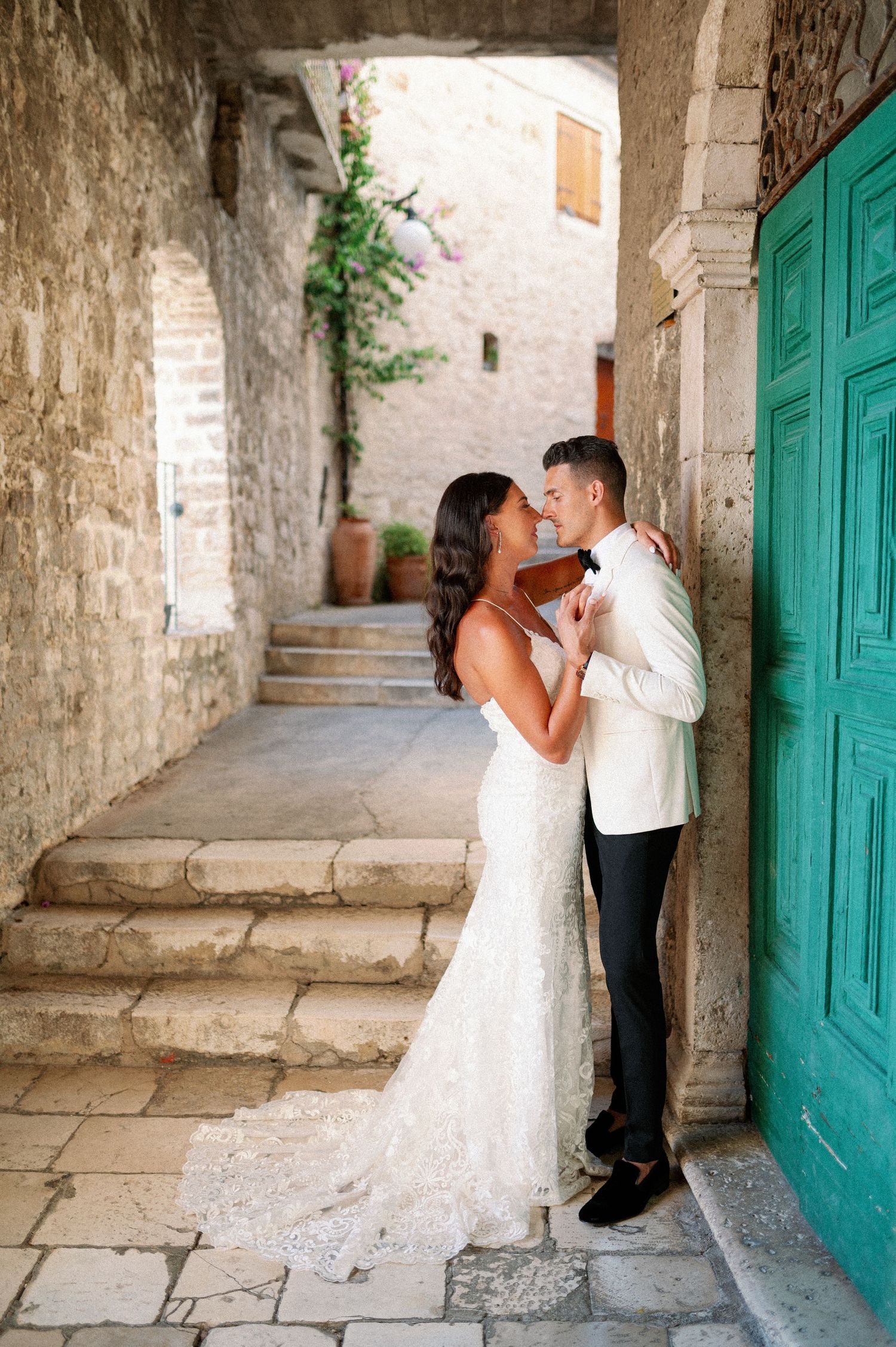 A romantic wedding couple embraces in a charming Mediterranean stone alleyway with a turquoise door and flowering vines.